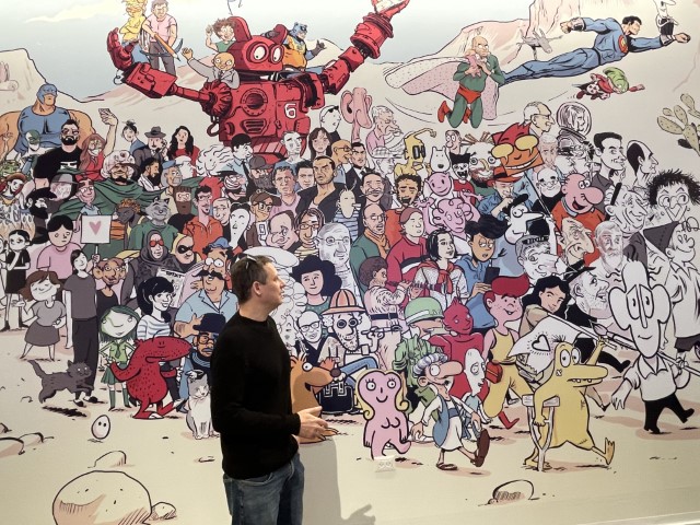 יונתן וקסמן לצד קיר הדיוקנאות של יוצרי הקריקטורה והקומיקס